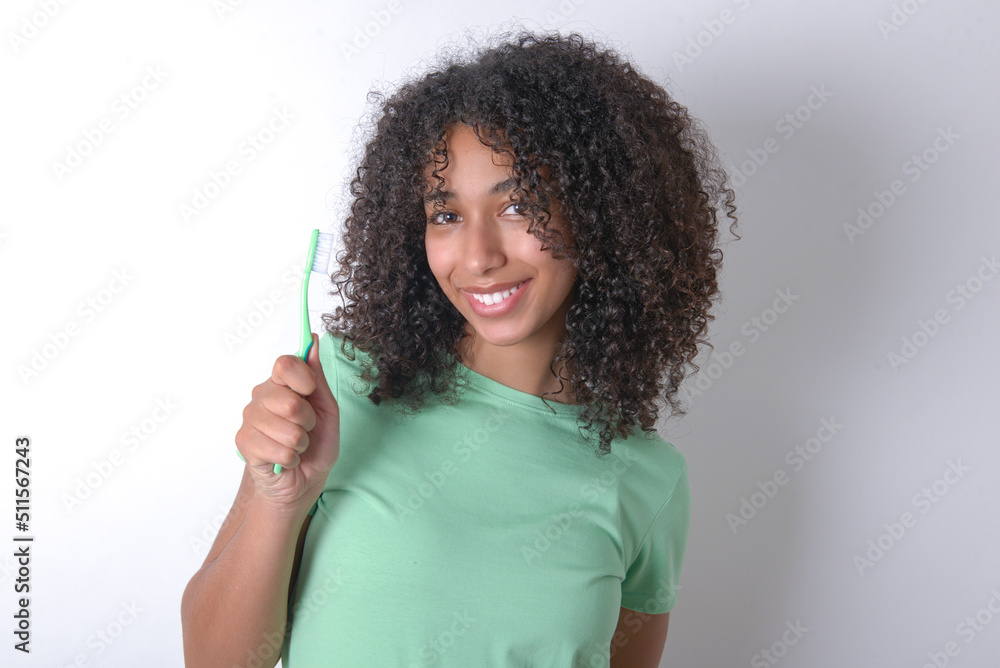 Young beautiful girl with afro hairstyle wearing green t-shirt over white background holding a toothbrush and smiling. Dental healthcare concept.