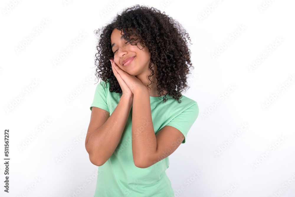Young beautiful girl with afro hairstyle wearing green t-shirt over white background leans on pressed palms closes eyes and has pleasant smile dreams about something