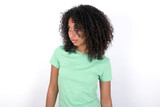 Young beautiful girl with afro hairstyle wearing green t-shirt over white background stares aside with wondered expression has speechless expression. Embarrassed model looks in surprise