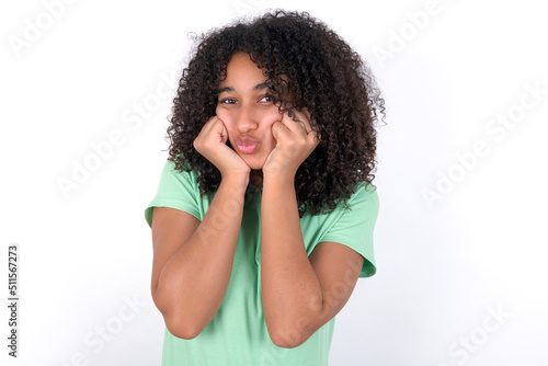 Young beautiful girl with afro hairstyle wearing green t-shirt over white background with surprised expression keeps hands under chin keeps lips folded makes funny grimace