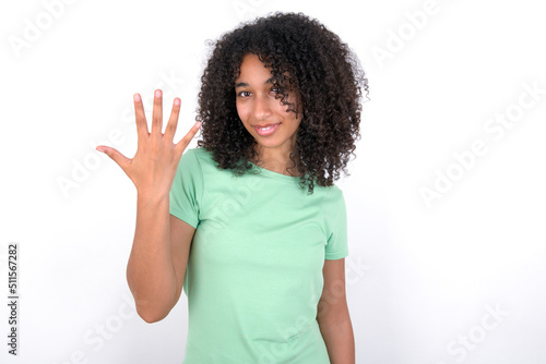 Young beautiful girl with afro hairstyle wearing green t-shirt over white background smiling and looking friendly, showing number five or fifth with hand forward, counting down