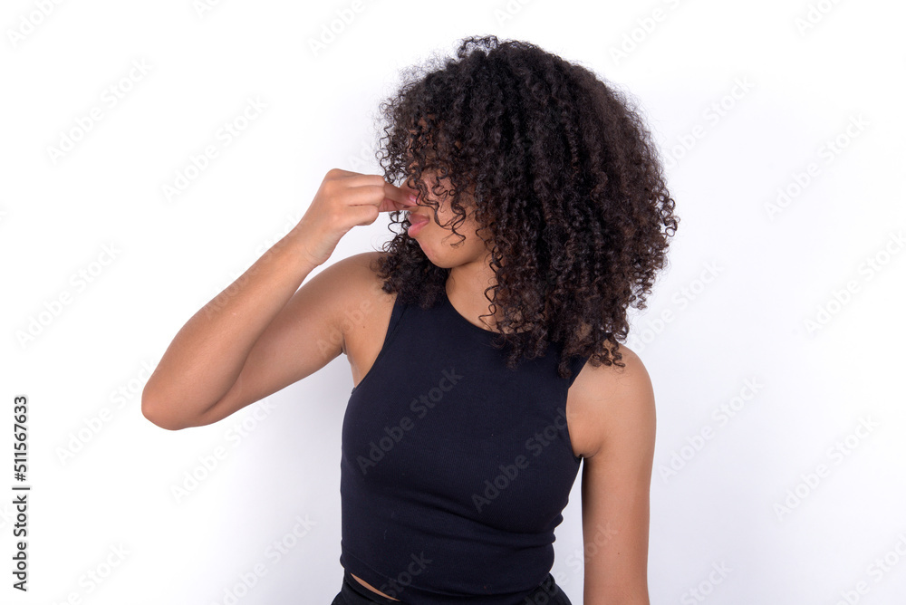 Young beautiful girl with afro hairstyle wearing black tank top over white background smelling something stinky and disgusting, intolerable smell, holding breath with fingers on nose. Bad smell Young beautiful girl with afro hairstyle wearing black tank top over white background smelling something stinky and disgusting, intolerable smell, holding breath with fingers on nose. Bad smell