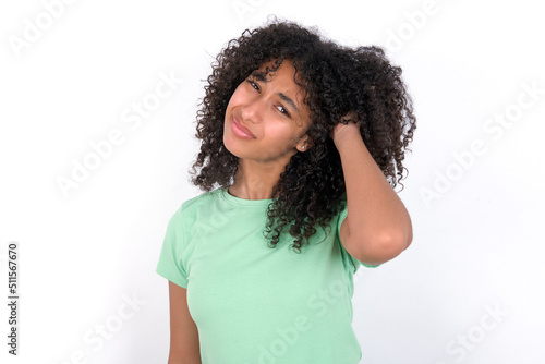 Young beautiful girl with afro hairstyle wearing green t-shirt over white background confuse and wonder about question. Uncertain with doubt, thinking with hand on head. Pensive concept.