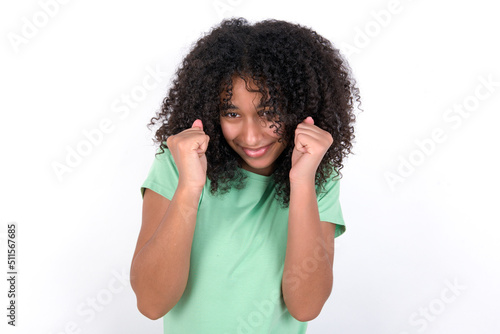 Young beautiful girl with afro hairstyle wearing green t-shirt over white background being excited for success with raised arms and closed eyes celebrating victory. Winner concept.