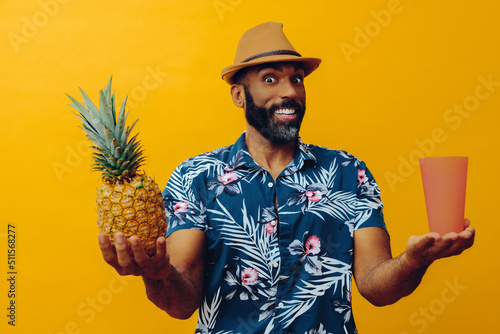 smiling mid adult african american man looking at camera wearing Hawaiian shirt and hat balancing pineapple and glass with hands to the sides studio 