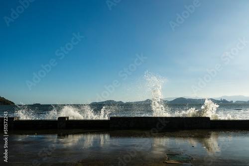 High tide waves crashing on the beach wall during winter with blue sky. City of Santos, Brazil. In the background on the right, Porchat Island.