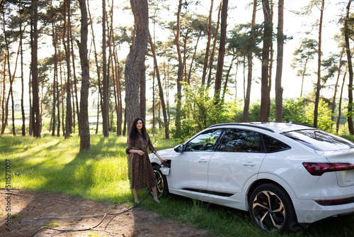 Beautiful young woman charges an electric car. Around the tree, sunset. Electric SUV