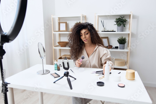 Professional makeup artist tanned handsome curly Latin beauty blogger in linen shirt tells how to sculpt body in home white interior. Copy space. Influencer record video review blog using smartphone