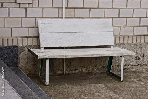 one empty wooden white bench stands on gray asphalt near the brick wall of the house in the yard