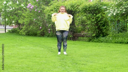 Positive red-haired curly overweight, adipose fat woman with excess weight in yellow hoodie doing sport exercises for neck in green park outdoor. Hold and rotate shoulders by hands in fresh air photo