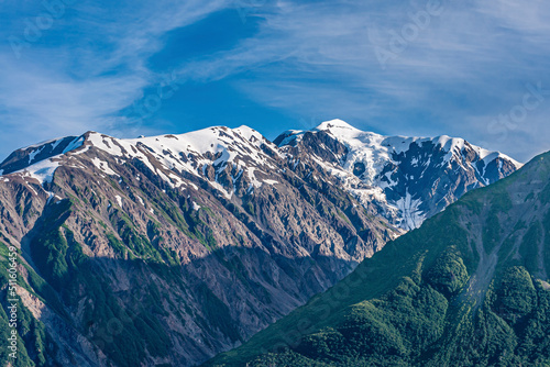 Disenchantment Bay, Alaska, USA - July 21, 2011: Closeup of snow topped gray mountain range under blue cloudscape. Green forested flank up front.