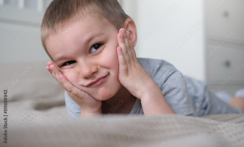 Close-up of a small happy caucasian boy in a gray t-shirt lies on the bed and smiles Close-up of a small happy caucasian boy in a gray t-shirt lies on the bed and smiles