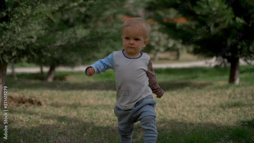 Blond toddler walks carefully on grass against lush green trees in spring park on sunny day. Adorable little boy experiences nature slow motion Blond toddler walks carefully on grass against lush green trees in spring park on sunny day. Adorable little boy experiences nature slow motion