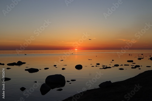 Fototapeta Naklejka Na Ścianę i Meble -  Sunset over calm Baltic sea, in Saulkrasti, Latvia. Sun shining from behind clouds, bright reflection on water, black shapes of boulders. Selective focus