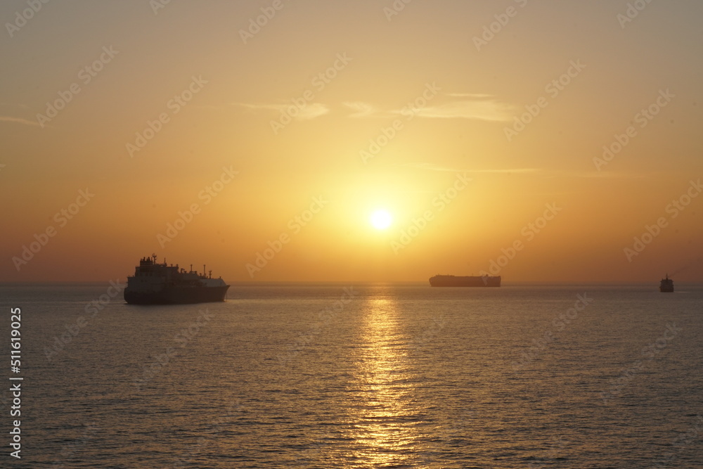 Cargo merchant vessels at anchor during sunset in Mediterranean Sea. The sky is displayed in varies harmonic shades of red, orange and yellow during calm weather in Sommer time. There is copy space.