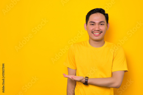 Smiling young Asian man in casual t-shirt holding copy space on palm for insert advertisement isolated on yellow background. People lifestyle concept
