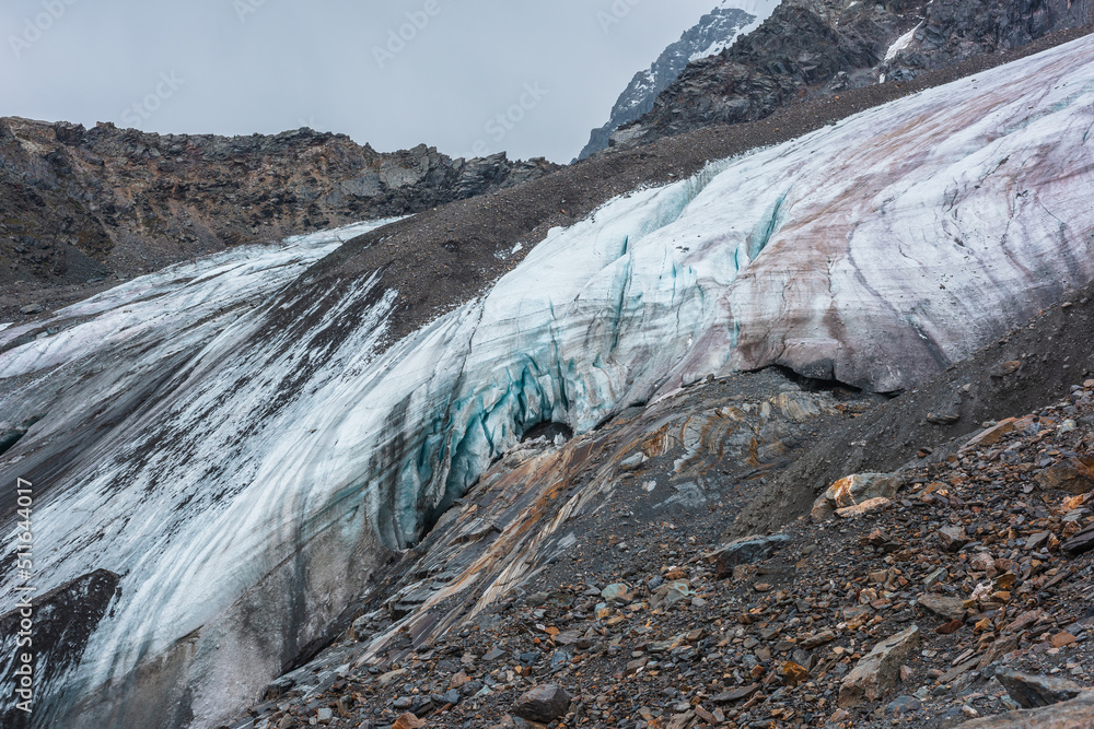 Awesome dramatic landscape with beautiful hanging glacier. Hole in dirty glacier near sharp mountain range. White blue glacial ice on smooth rocks of orange color. Cavity in ice cornice in center. Awesome dramatic landscape with beautiful hanging glacier. Hole in dirty glacier near sharp mountain range. White blue glacial ice on smooth rocks of orange color. Cavity in ice cornice in center.