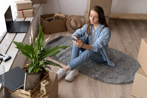 Beautiful girl is using a smartphone, looking at camera and smiling while sitting on the floor near packed boxes ready to move, young woman texting while moving to new apartment © mtrlin