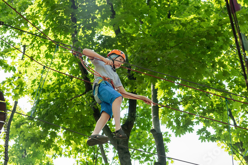Rope park. A boy teenager in a helmet walks on suspended rope ladders. Carabiners and safety straps. Safety. Summer activity. Sport. Children's playground in nature in the forest.