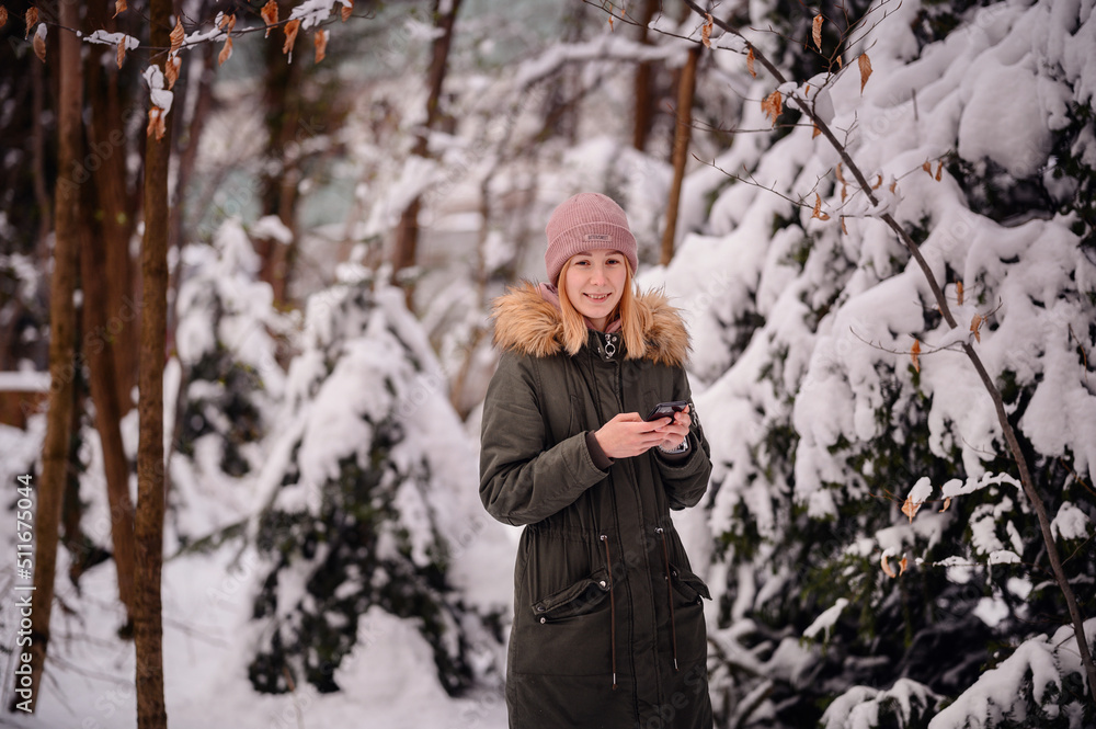 Young blonde girl in winter outfit stands in snowy park using smartphone Young blonde girl in winter outfit stands in snowy park using smartphone