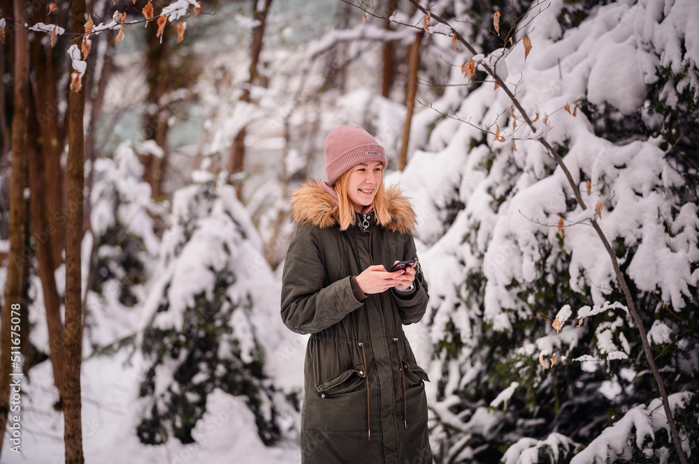Young blonde girl in winter outfit stands in snowy park using smartphone Young blonde girl in winter outfit stands in snowy park using smartphone