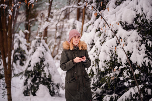 Young blonde girl in winter outfit stands in snowy park using smartphone