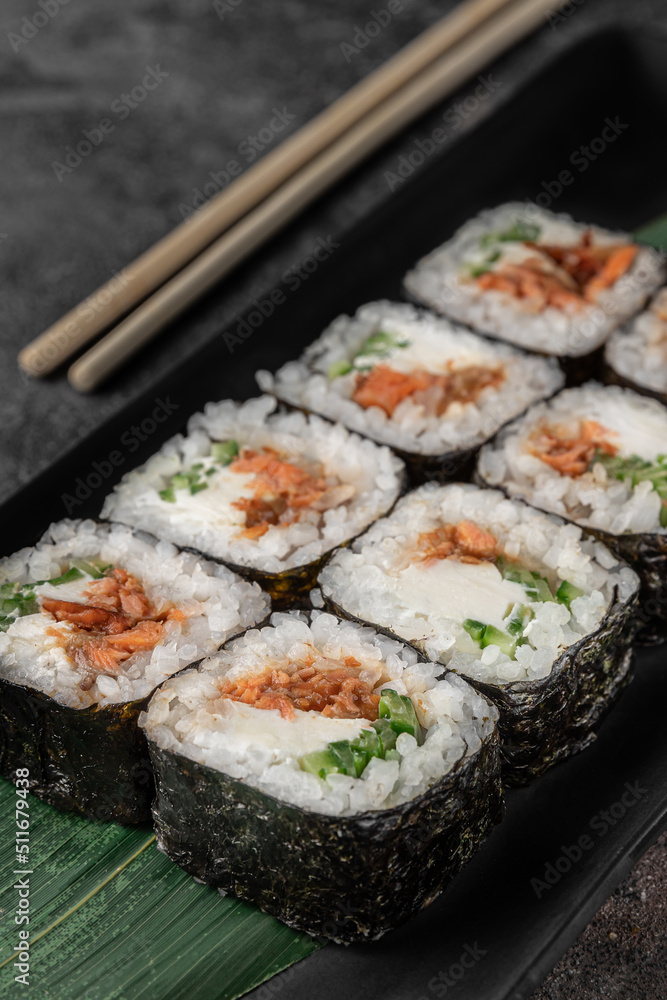 set of square maki rolls with fried salmon, curd cheese and green bamboo leaf in a black ceramic plate with chopstick on a dark gray textured background, side view, close-up set of square maki rolls with fried salmon, curd cheese and green bamboo leaf in a black ceramic plate with chopstick on a dark gray textured background, side view, close-up