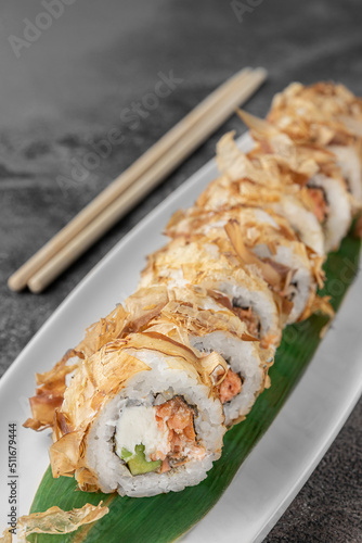 set of bonito rolls with grilled salmon, avocado, tuna shavings and green bamboo leaf in a white ceramic plate with chopstick on a dark gray textured background, side view, close-up