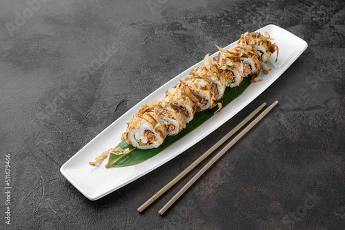 set of bonito rolls with grilled salmon, avocado, tuna shavings and green bamboo leaf in a white ceramic plate with chopstick on a dark gray textured background, side view