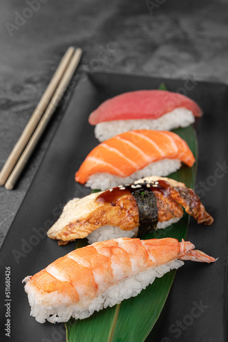 set of nigiri with tiger shrimp, tomago, salmon, tuna and green bamboo leaf in a black ceramic plate with chopstick on a dark gray textured background, side view, close-up