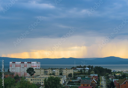 Residential houses and buildings against the backdrop of a lake and clouds and rain over the mountains in the distance against a yellow sky