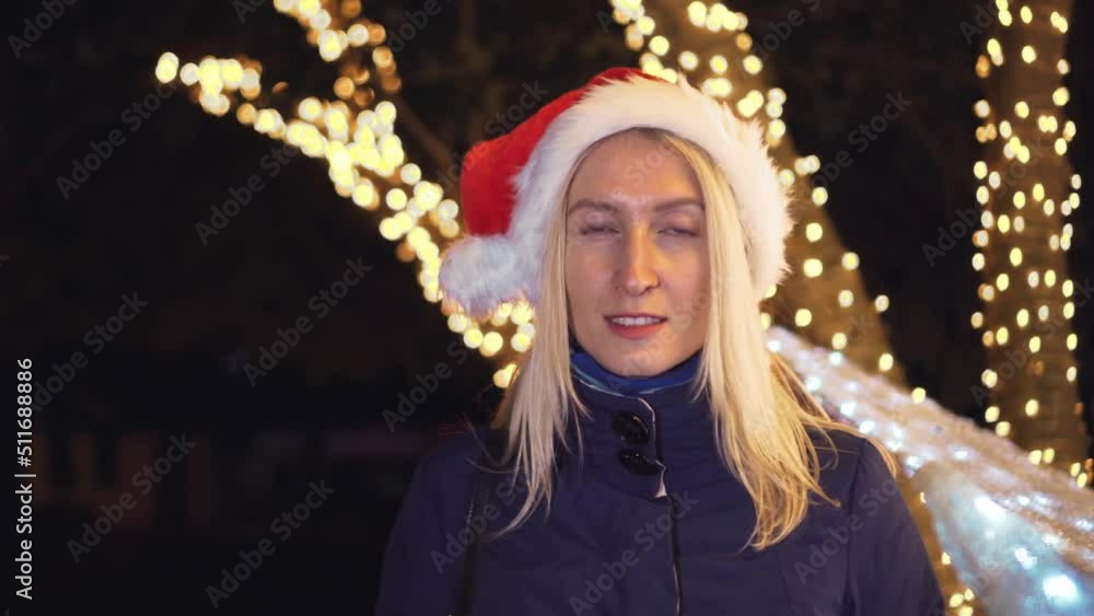 Portrait of a woman with makeup, smiling at the camera and playing in a Santa hat against background of city lights at night or in the evening. She smiles, rejoices celebrating New year or Christmas. Portrait of a woman with makeup, smiling at the camera and playing in a Santa hat against background of city lights at night or in the evening. She smiles, rejoices celebrating New year or Christmas.