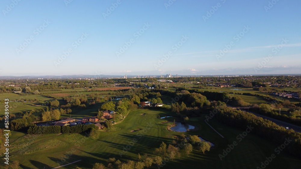 Aerial view looking down onto a golf course surrounded by green fields with the Manchester skyline in the distance. 
