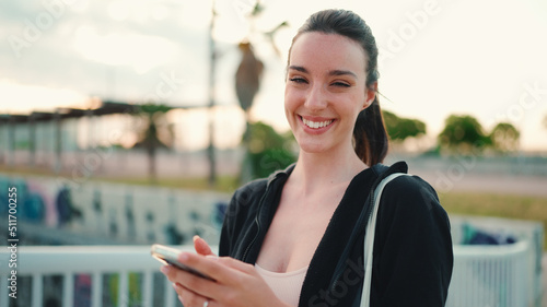Young woman with freckles and long ponytail wearing black hoodie and beige sports top uses mobile phone and listens to music while standing on bridge modern city background