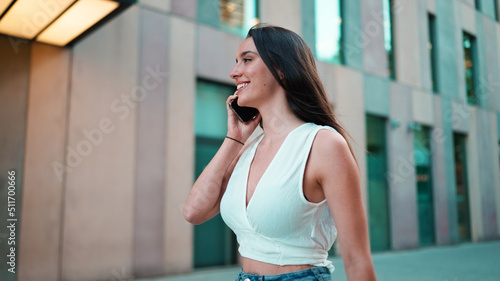 Beautiful woman with freckles and dark loose hair wearing white top is walking down the street with smartphone in her hands. Cute girl talking on mobile phone on modern city background