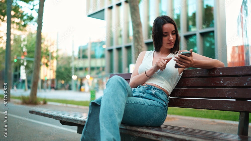 Beautiful woman with freckles and dark loose hair wearing white top sits on bench with phone in her hands. Cute smiling girl watching photo, video on mobile phone modern city background