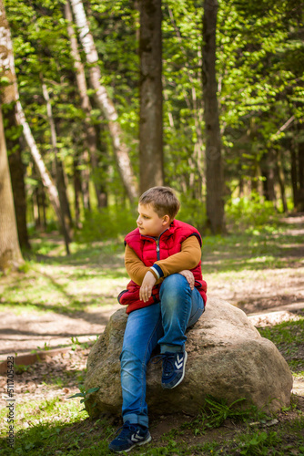 Portrait of a young man in a red tank top in the forest in spring. Walk through the green park in the fresh air. The magical light from the sun's rays falls behind the boy. © Alina Lebed