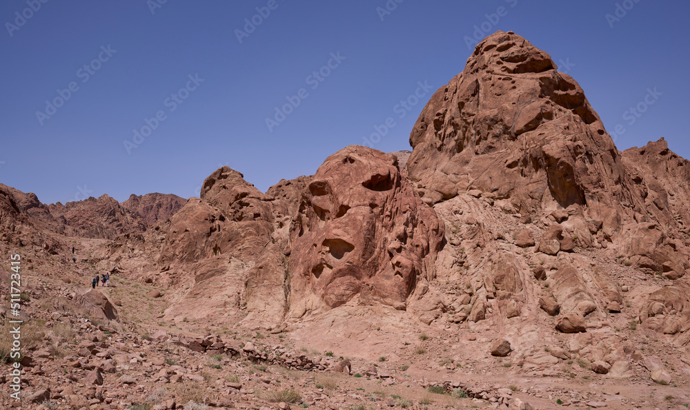 Group of hikers on a hiking trail in Southern Sinai, Egypt. Panoramic view on surrounding red mountains, huge boulders and rocks of amazing shapes. Face shaped rock formation. 