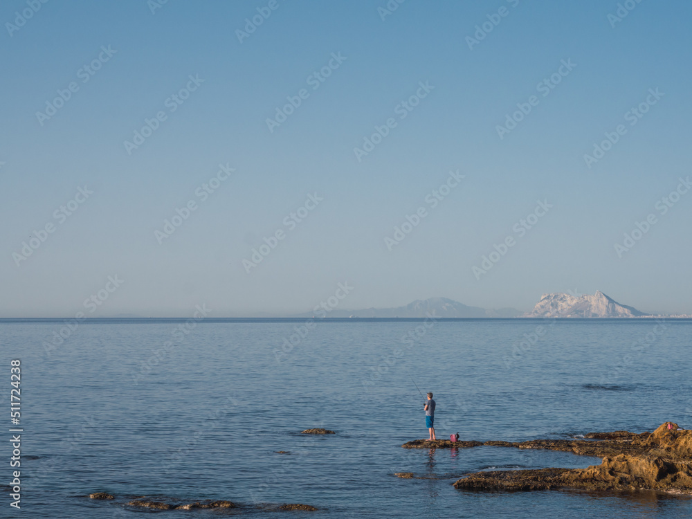 Man fishing in the Mediterranean Sea and with a blue sky with the Rock of Gibraltar and the Altas Mountain (Africa) in the background. You see another continent