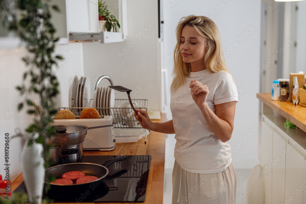Pretty young female standing with spatula at kitchen and frying burger meat. Pretty young female standing with spatula at kitchen and frying burger meat.