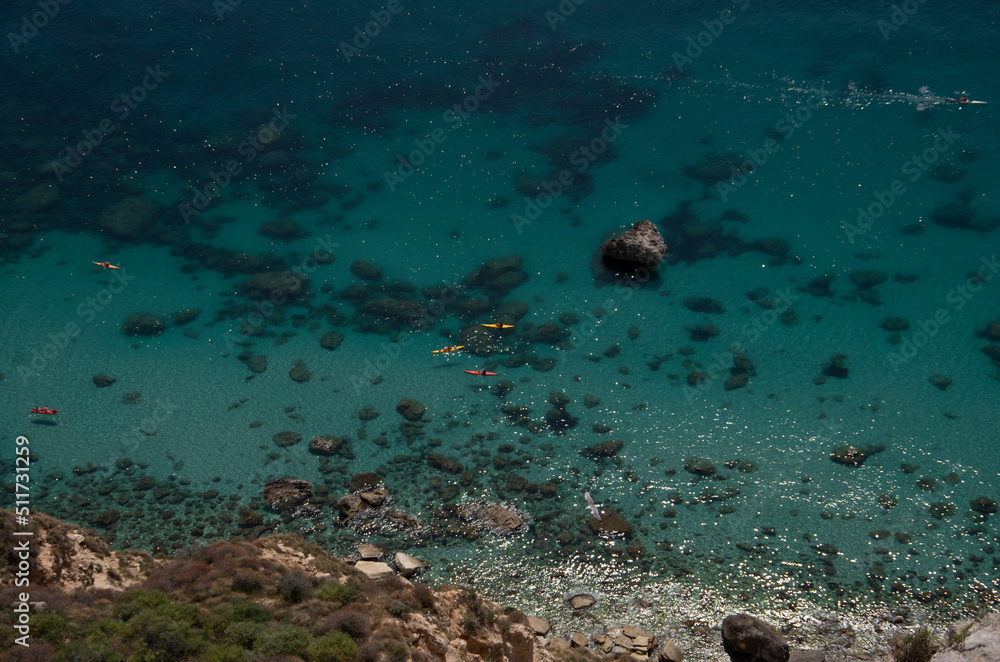 Crystal Sardinian water from a top view, with people doing kaykaking with red and yellow boats floating on the surface. Clear and visible seabed, with fine sand and small rocks. 