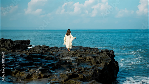 A young European girl with long black hair and a white robe walks to Water blow with cliffs and strong waves of seawater with clouds and blue sea