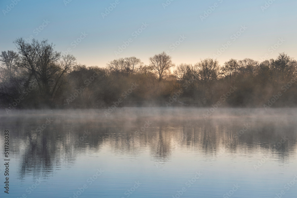 Beautiful landscape image of sunrise mist on urban lake with sun beams streaming through trees lighting up water surface