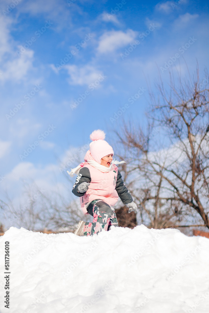Child rides on snow slide. Little girl in winter warm suit plays in fresh air and enjoys sunny day in winter, she lies in snow and plays snowballs.