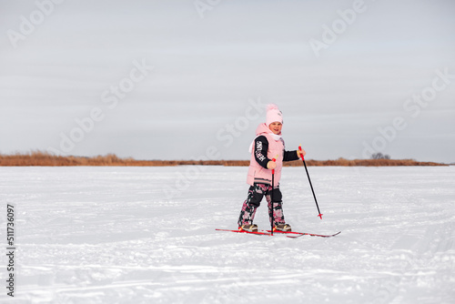 Child learns to ski. Little girl in pink warm suit skiing in snow on frosty winter day, side view, snow background. 