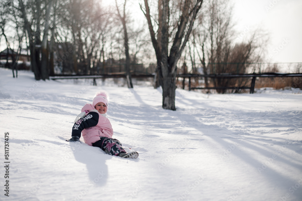 Child plays in park in winter. Little girl rides snow slide and enjoys warm sunny day on winter holidays.