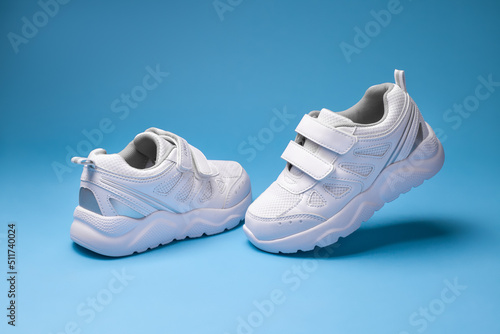front view white child sneakers with velcro fasteners  one sneaker flying in the air and the other standing on the floor isolated on a blue background.