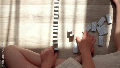 Close-up top view of little caucasian girl child builds lines with small dominoes sitting alone on warm floor at home. Concept domino effect in business, education, development fine motor skills photo