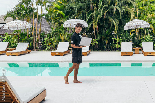young man works on silver laptop computer remotely near beautiful pool © Alexander White