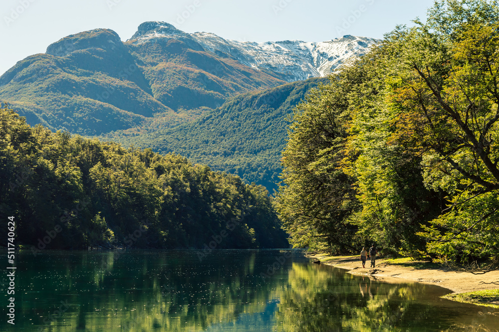 Couple walking along the coast of a river that reflects with a mountain range in the background. Arrayanes River, Los Alerces National Park.